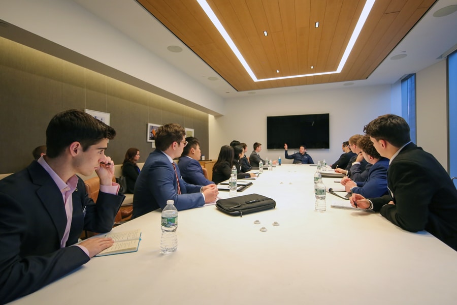 A group of business students in formal attire attentively listens to a speaker at the head of a long conference table in a modern boardroom.