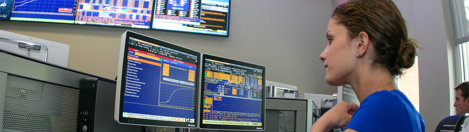 Student focused on analyzing financial data on multiple monitors in a modern trading room environment.