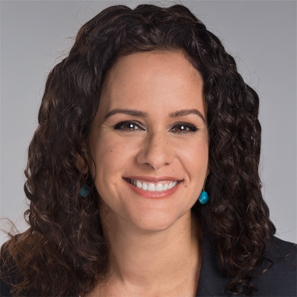 Professional headshot of Yessi Scheker Izquierdo, a woman with long, curly dark hair, smiling confidently, wearing turquoise earrings and a black blazer, against a light gray background.