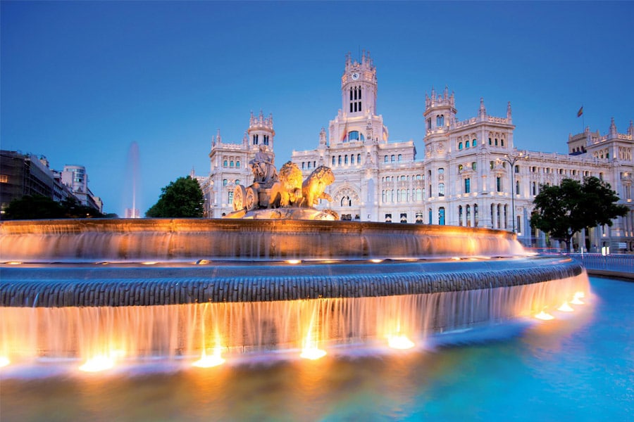 Illuminated fountain at Cibeles Square in Madrid, with water cascading over tiers with the Palacio de Cibeles peering from behind in the twilight sky.