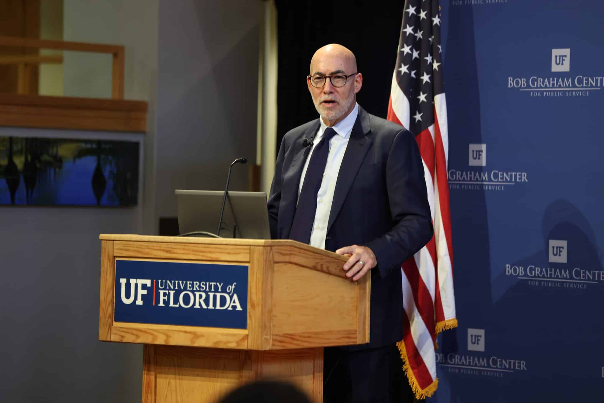 Peter Goodman stands at a podium with University of Florida written on the front, while speaking. He wears a suit and stands in front of an American flag. 