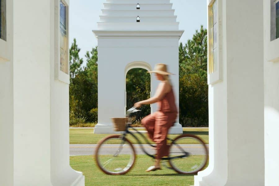 Through a white structure opening a woman can be seen riding a bicycle on the grass as the sun shines with another white structure in the background.