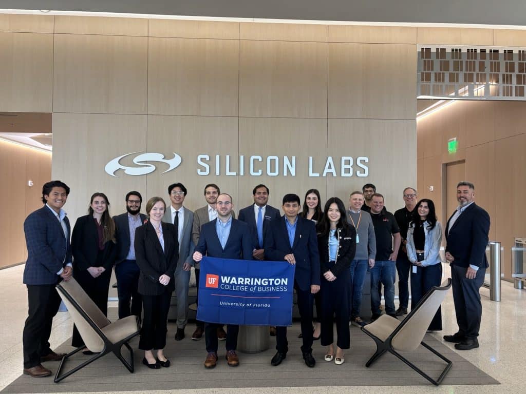 Students in Mike Carillo's MAN6930 Fall 2025 Pose in front of a Silicon Labs sign, holding a Warrington College of Business Flag.