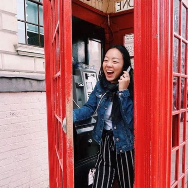 A young woman laughs as she holds a phone to her ear inside a red London phone booth