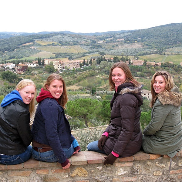 Four young women seated on a stone wall turn to smile at the camera with rolling hills, some buildings and farmland in the background