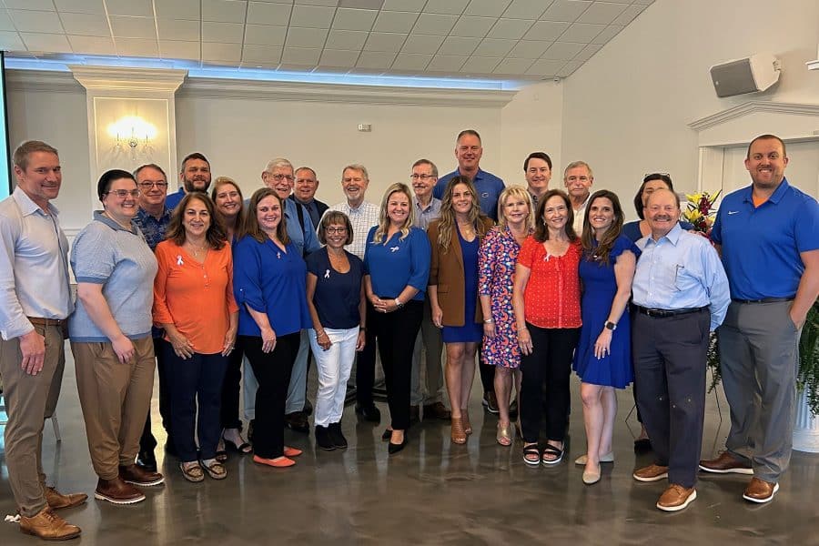 A group of about 20 people, many wearing orange and blue, representing past and present partners at James Moore & Co's office.