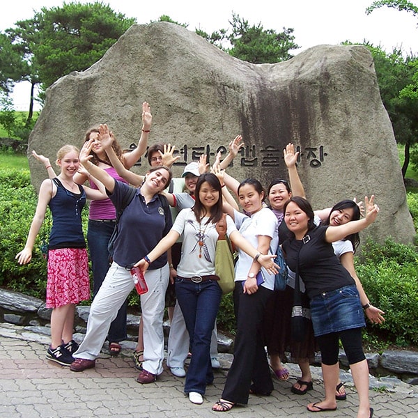 Students pose with arms outstretched in front of a large stone that has Korean writing on it.