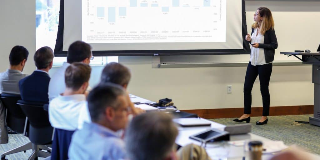 A woman stands at the front of a room next to a projected slideshow with a bar graph showing as individuals seated at a long table focus on the presentation.