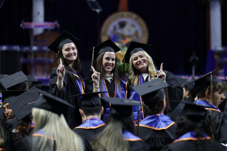 Three female students wearing graduation regalia stand among a group of seated graduates while holding up their pointer finger, indicating #1.
