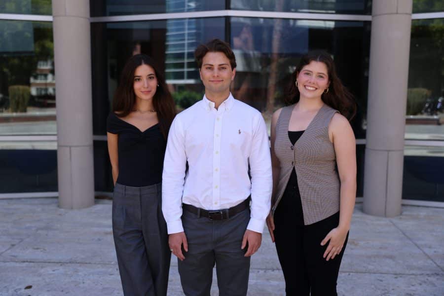 Matthew Smith, Cecelia (Cece) Duncan and Alejandra Sammataro pose together in front of the glass windows for a photo outside of Hough Hall.