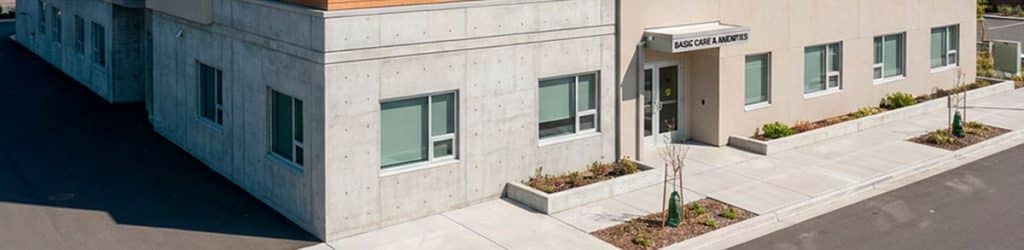 A building front with few windows, cement exterior and minimal landscaping. It says base care amenities above the door.