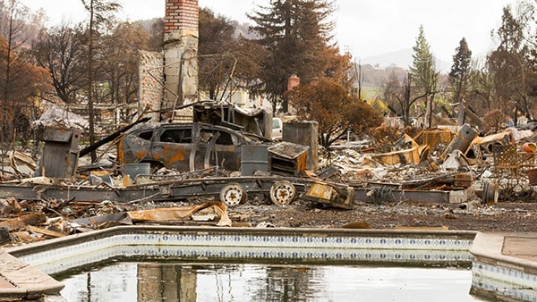 An in-ground pool surrounded by debris from a home, trailer and vehicle that seem to have burned up. A brick chimney remains standing amidst the destruction.