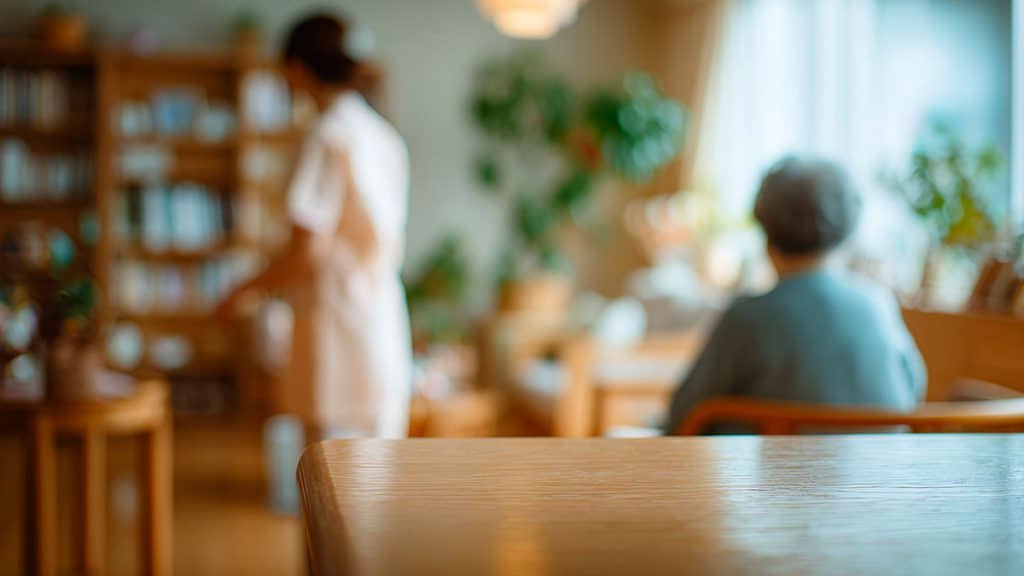 Focal point on a table corner while in the blurred background, an elderly woman is seated facing a younger person who is standing or walking in a common area with seating, tables, books and plants.
