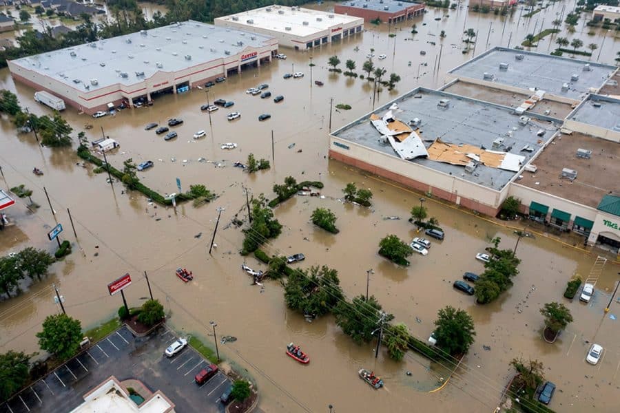An aerial view of a flooded shopping area with big box stores. Vehicles are parked in standing water while rescue rafts pass by and storm damage can be seen on one of the roofs.