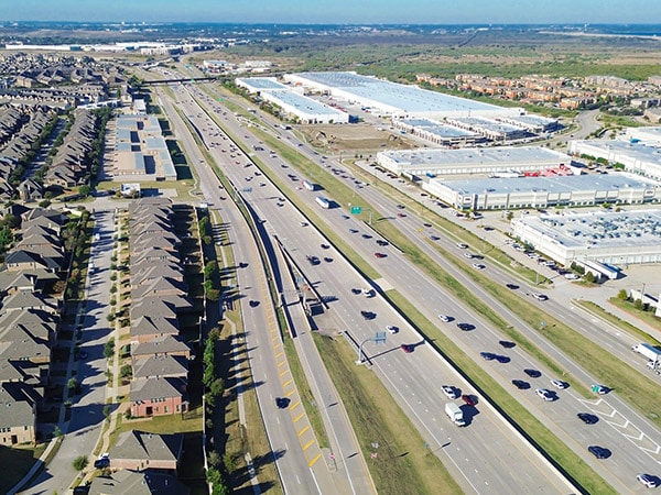 Cars and trucks traveling on an interstate through a Texas city with homes on one side and large factories, warehouses or shipping centers on the other.