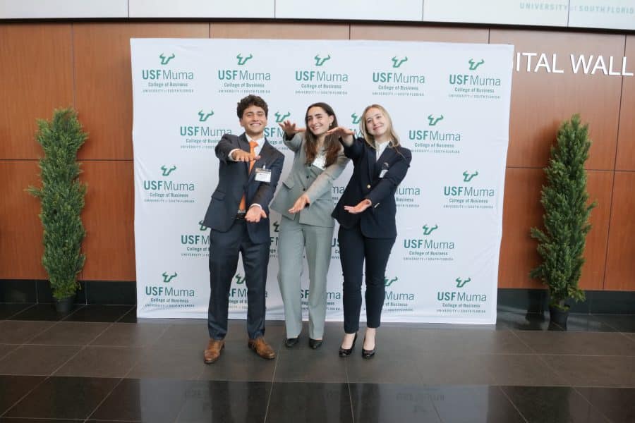 Sophia Miele, Catherine McGarry and Daniel Khani standing in front of a USF Muma sign smiling and doing the Gator chomp