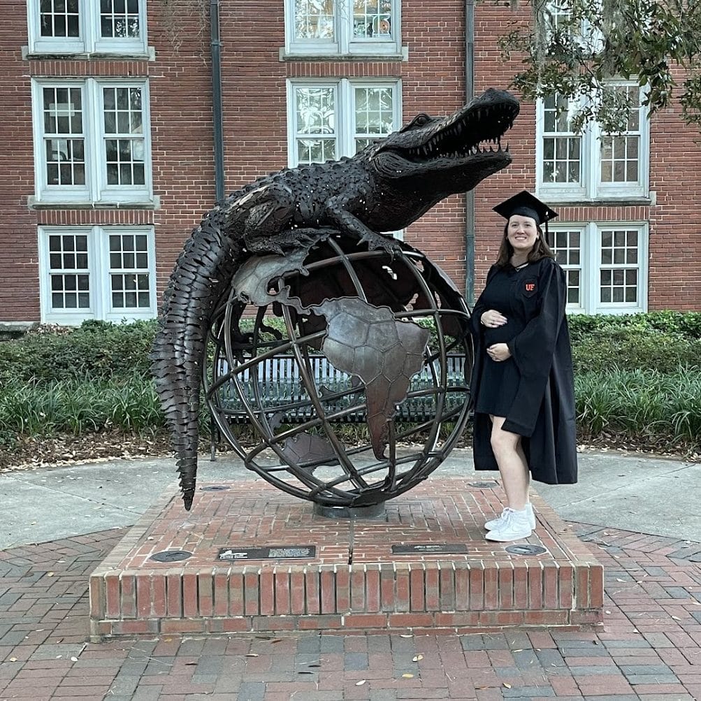 MBA student Katie Mazzola in a graduation cap and gown pregnant, holding her stomach in front of the heavener gator globe statue