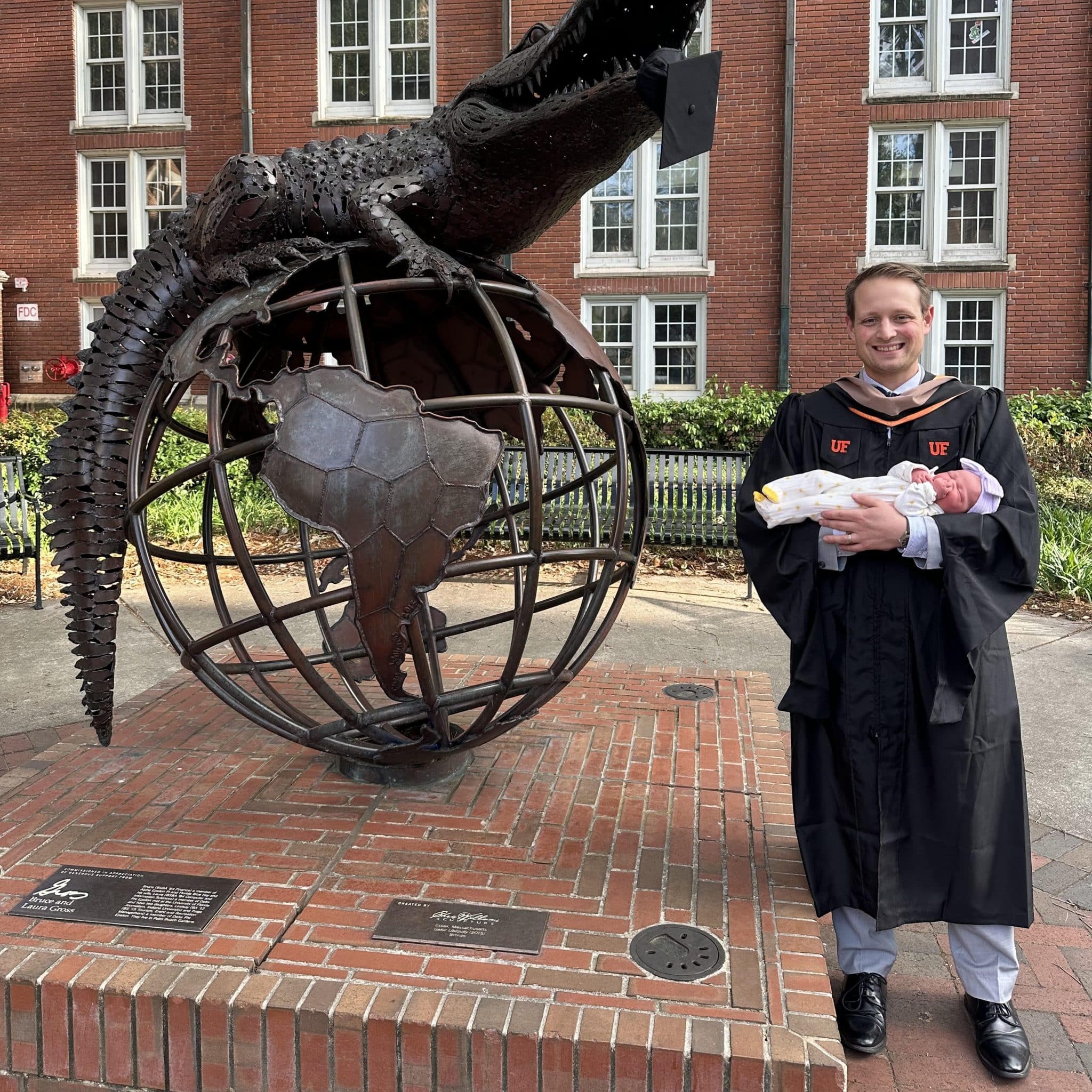 MBA student Matthew Brown in a graduation gown holding a baby girl in front of the heavener gator globe statue with his cap in the gator's mouth