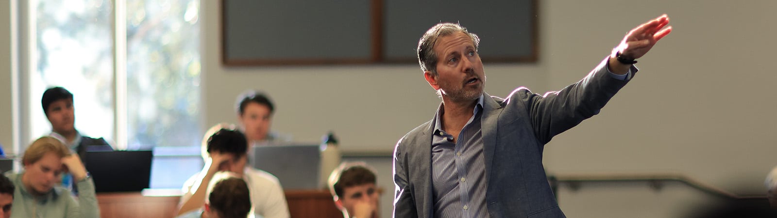 A professor gestures to the front of the room as students behind him listen to what he is teaching.