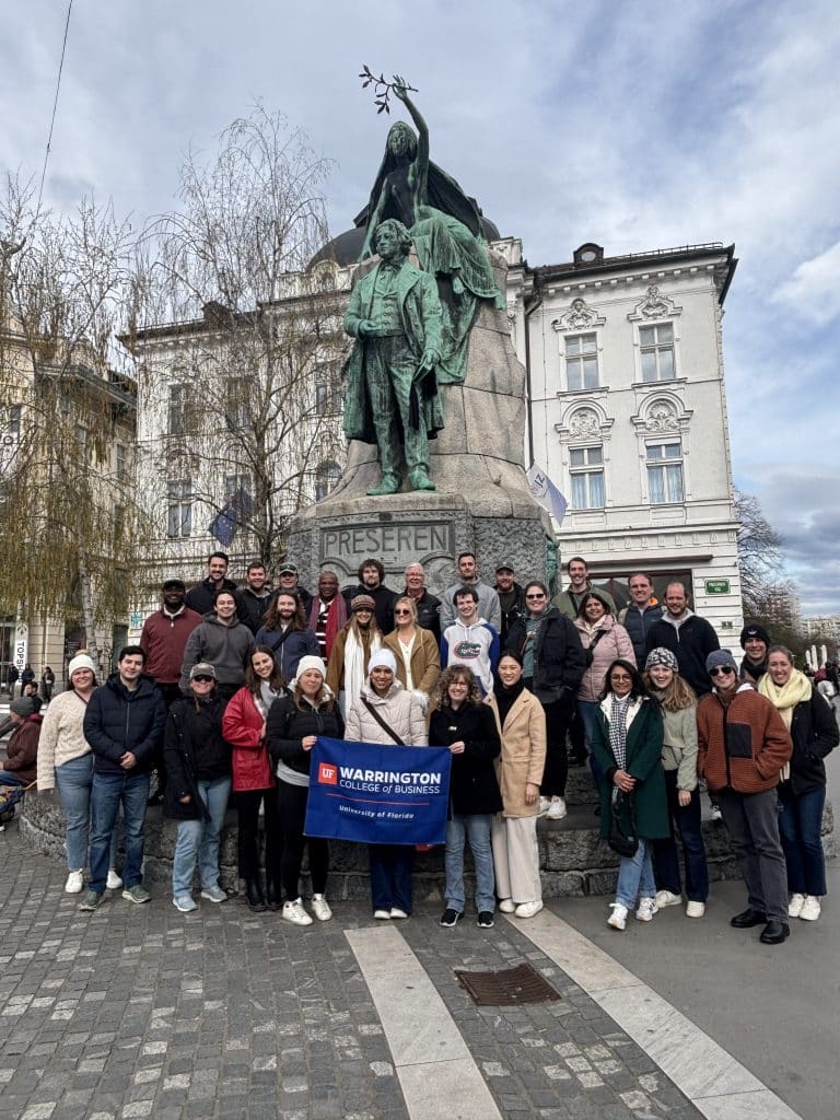 MBA students pose for a group photo in front of a statue on the Croatia Global Immersion Experience. One student holds a banner that says "Warrington College of Business, University of Florida."