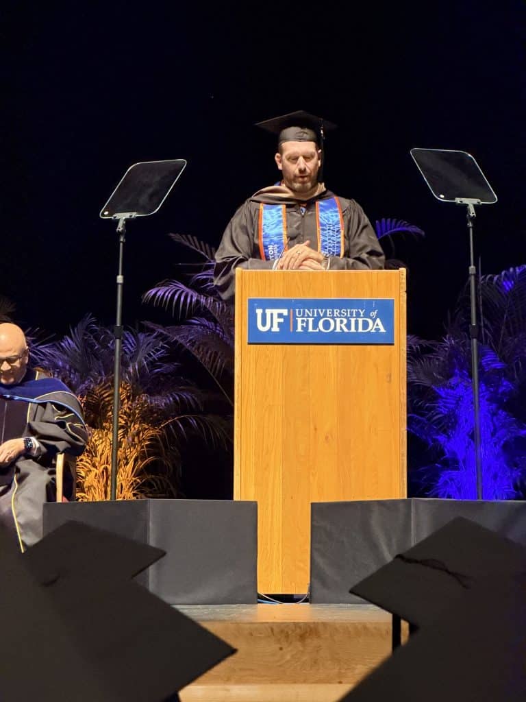 Chris Taylor in a cap and gown standing on a stage with a podium giving a speech