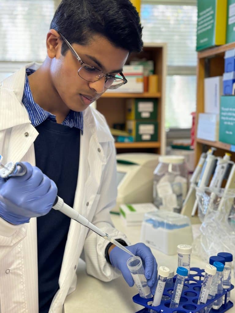 Abhith Kasala in a science lab with glasses, gloves and a lab coat holding a pipette and beaker