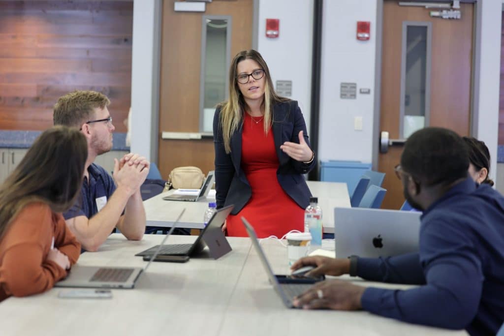 Kat Haney speaking to four students sitting at a table looking at her