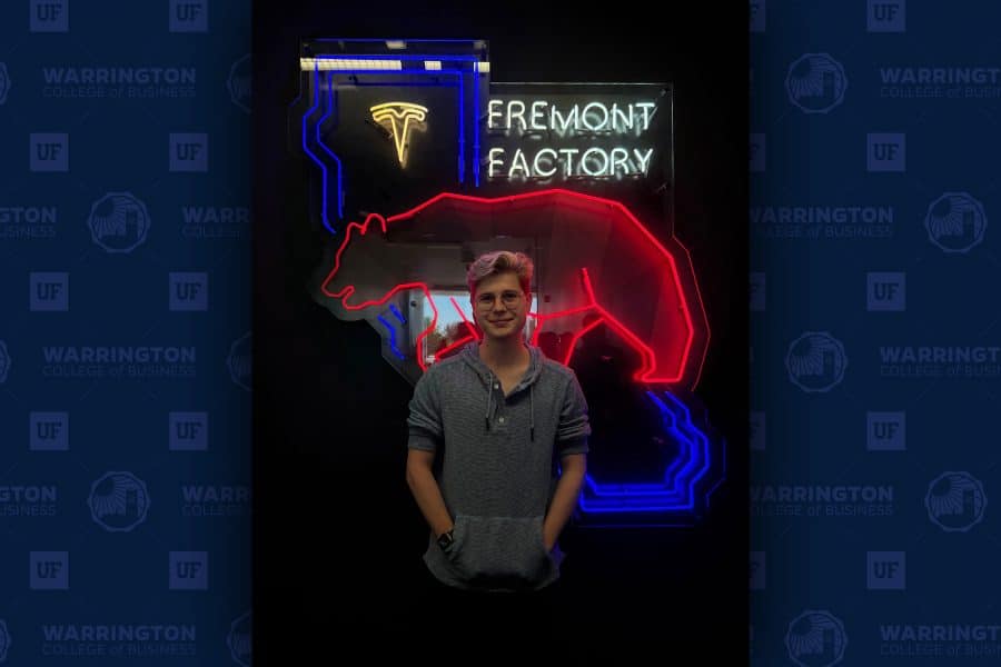 Cole McKinney stands in front of a glowing neon sign that says "Fremont Factory." Also in neon appears the Tesla logo, the outline of the state of California, and the Tesla Cyberbear logo.