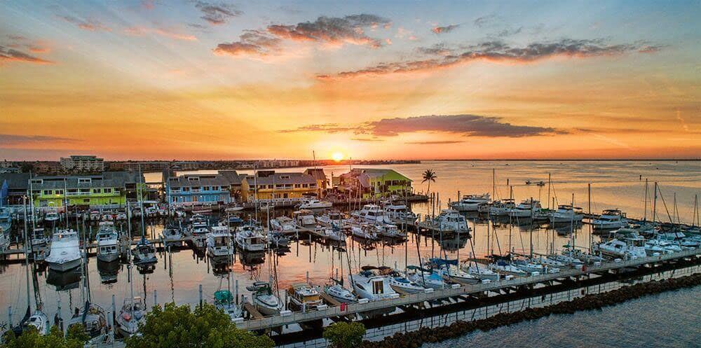 Aerial view of a waterfront marina with several boats against an orange sunset. 