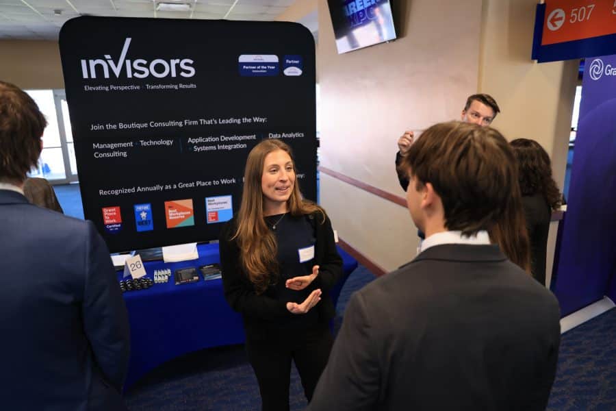 A recruiter stands in front of a sign with the company's logo while speaking with a student at a career fair.