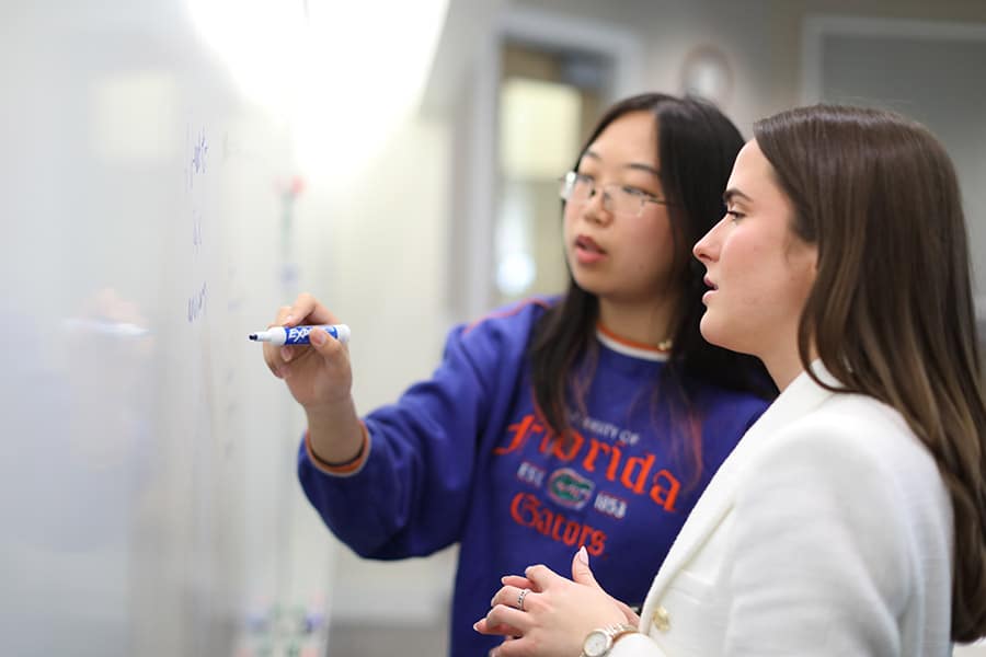 Two students stand at a white board as one holds a blue marker and writes, they both focus on their task.